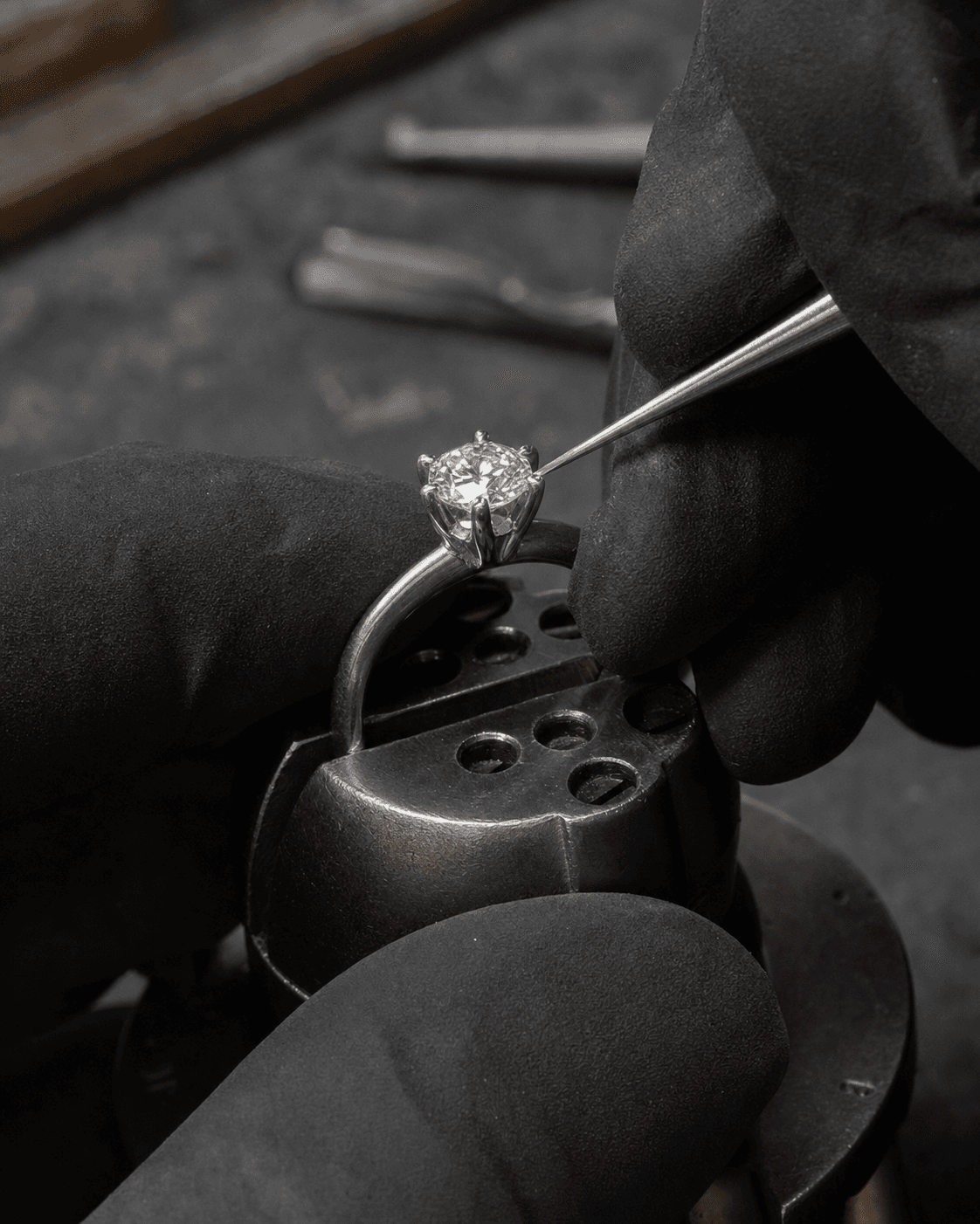 Jeweler’s gloved hands using a precision tool to adjust prongs on a diamond solitaire held in a bench vise